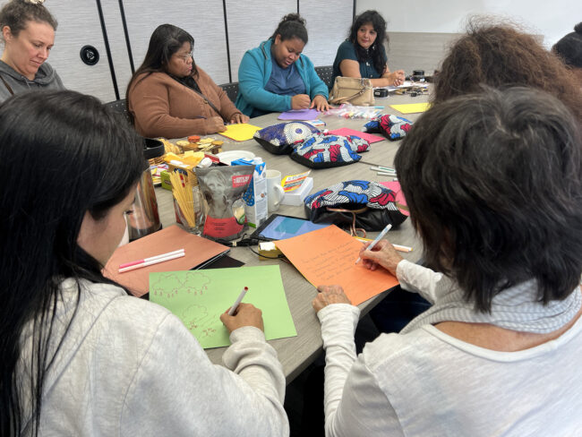 Plusieurs femmes sont assises en cercle autour d'une table remplie de coussins vibrotactiles, de tablettes tactiles, de feutres, de papier et de collations. Elles font des dessins de lieux qu'elles trouvent reposants.