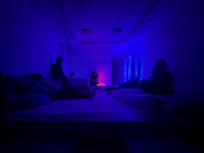 A dimly lit room with blue and purple ambient lighting where silhouettes of people are relaxing and resting. Une pièce faiblement éclairée avec des tons de bleu et de violet où l’on devine des silhouettes de personnes qui se détendent et se reposent.