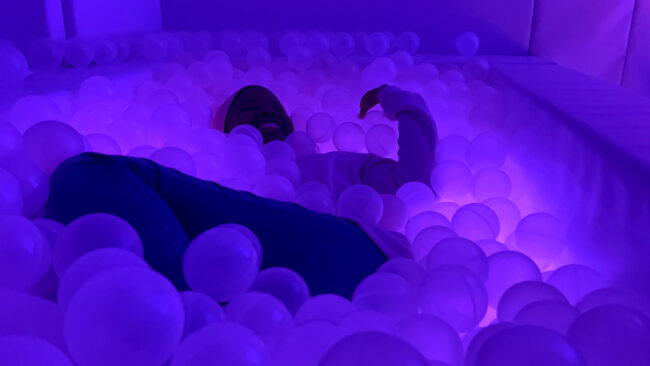 The silhouette of a person smiling while immersed in a ball pit with white spheres, illuminated by purple lighting in an indoor sensory environment. Silhouette d'une personne souriante immergée dans une piscine à balles blanches, éclairée de lumière violette, dans une salle multisensorielle.
