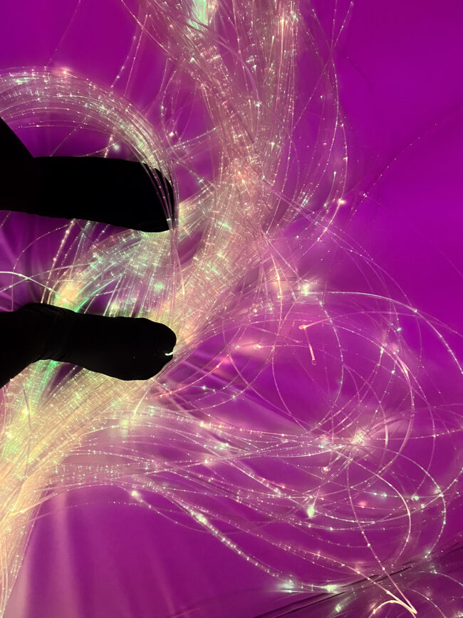 Feet resting on fiber optic light strands that create flowing, luminous patterns against a purple-pink background, resembling aurora-like light effects. Des pieds reposent sur des brins de fibre optique lumineuse qui créent des motifs fluides sur un fond violet-rose, effets de lumière rappelant l’aurore.