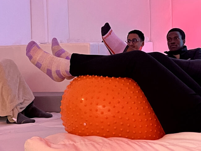 Two people sitting on a white couch in a softly lit room with pink and purple ambient lighting, using an orange textured therapy ball for sensory activities. Deux personnes, assises sur un canapé blanc dans une pièce doucement éclairée de rose et de violet, utilisent un ballon de thérapie texturé orange pour des activités sensorielles.