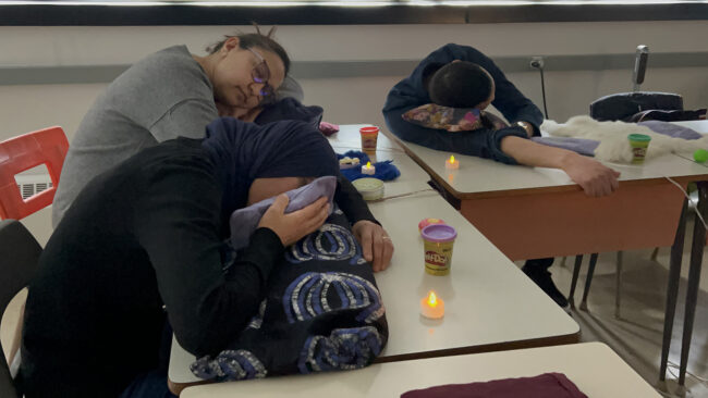 Students lying down at their desks in a classroom, participating in a restful listening activity, with small candles providing gentle lighting. Des élèves, allongés à leur bureau dans une salle de classe, participent à une activité d'écoute reposante, avec de petites bougies offrant un éclairage doux.