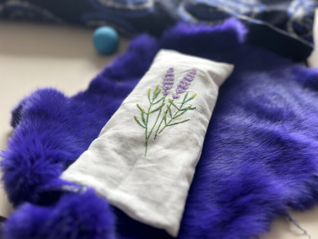soft purple fur or fabric. Un coussin pour les yeux en tissu blanc brodé de fleurs de lavande repose sur une douce fourrure violette.