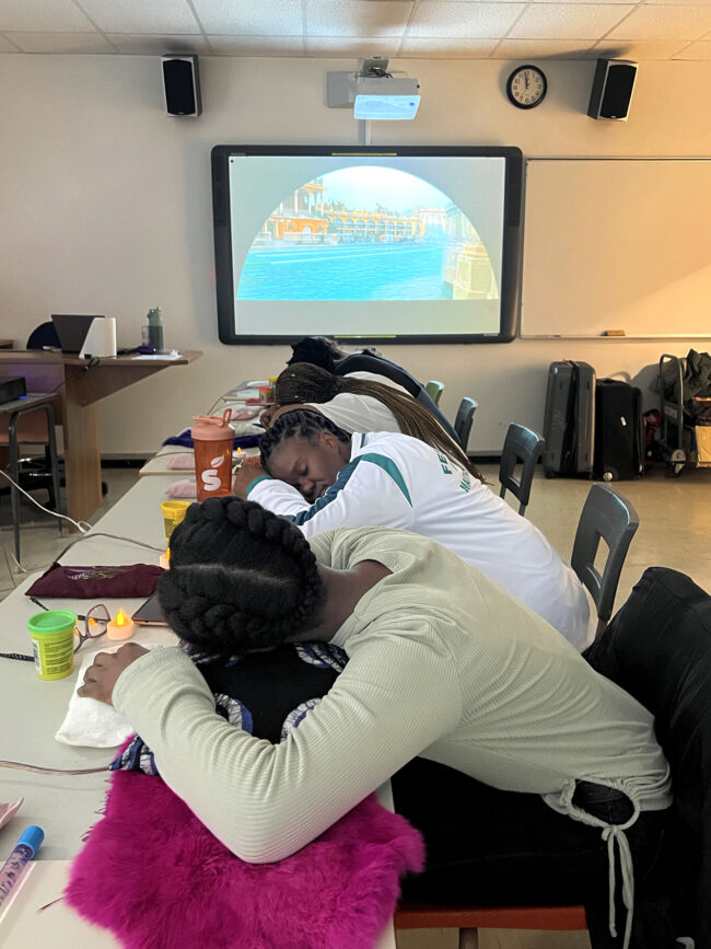 Students resting at desks in a classroom with a projected image of a bathhouse on the wall, creating a calming atmosphere. Des élèves se reposent à leur bureau dans une salle de classe. Une vidéo de bain public est projetée sur le mur, créant une atmosphère apaisante.