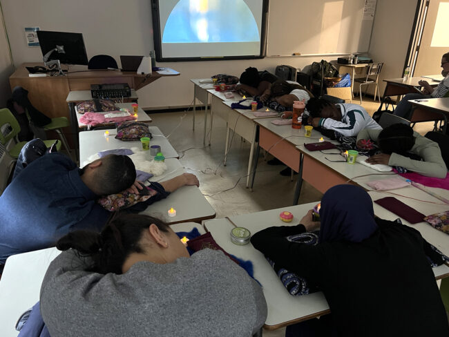 A classroom full of students lying down at their desks during a guided listening and rest session, with a peaceful projected image visible on the wall. Une salle de classe pleine d'élèves allongés à leur bureau pendant une séance d'écoute guidée et de repos. Une image paisible est projetée sur le mur.